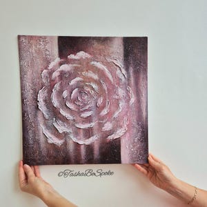 May include: Square canvas art with a textured rose design in pink and white against a dark brown and pink backdrop. The artwork is held by hands and has the text "TashasBeSpoke" at the bottom.