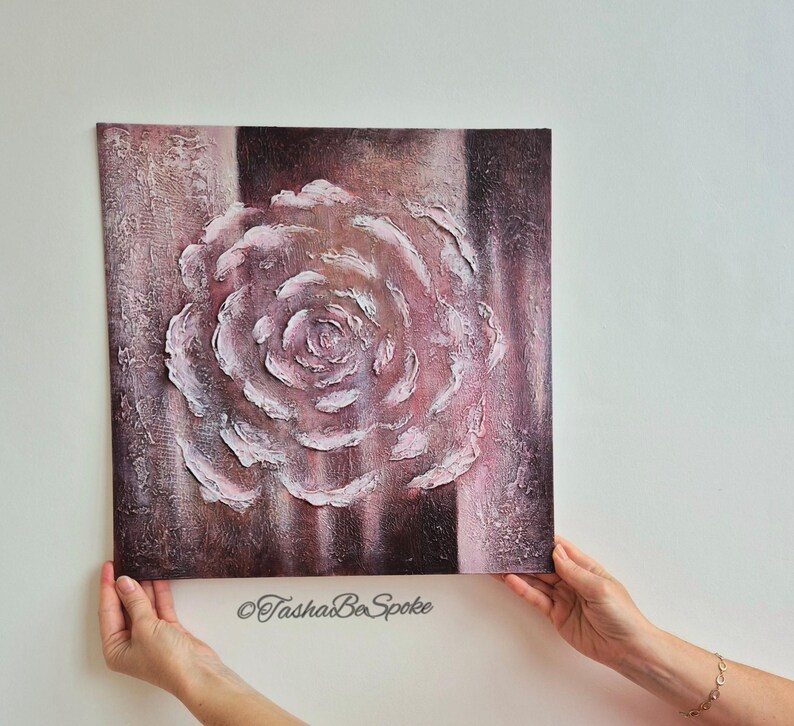 May include: Square canvas art with a textured rose design in pink and white against a dark brown and pink backdrop. The artwork is held by hands and has the text "TashasBeSpoke" at the bottom.