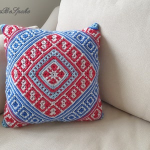 Op de afbeelding: Vierkant gehaakt kussen met een geometrisch patroon in rood, wit en blauw. Het kussen heeft een centraal ruitontwerp omgeven door herhalende vierkanten en ruitvormen. Het kussen ligt op een cr&egrave;mekleurige bank.