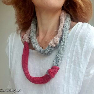 May include: A knitted necklace in shades of gray, pink, and red. The necklace is knotted in several places, with a bright red section at the bottom. The design is handmade and features a unique, textured appearance.