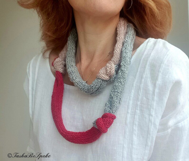 May include: A knitted necklace in shades of gray, pink, and red. The necklace is knotted in several places, with a bright red section at the bottom. The design is handmade and features a unique, textured appearance.