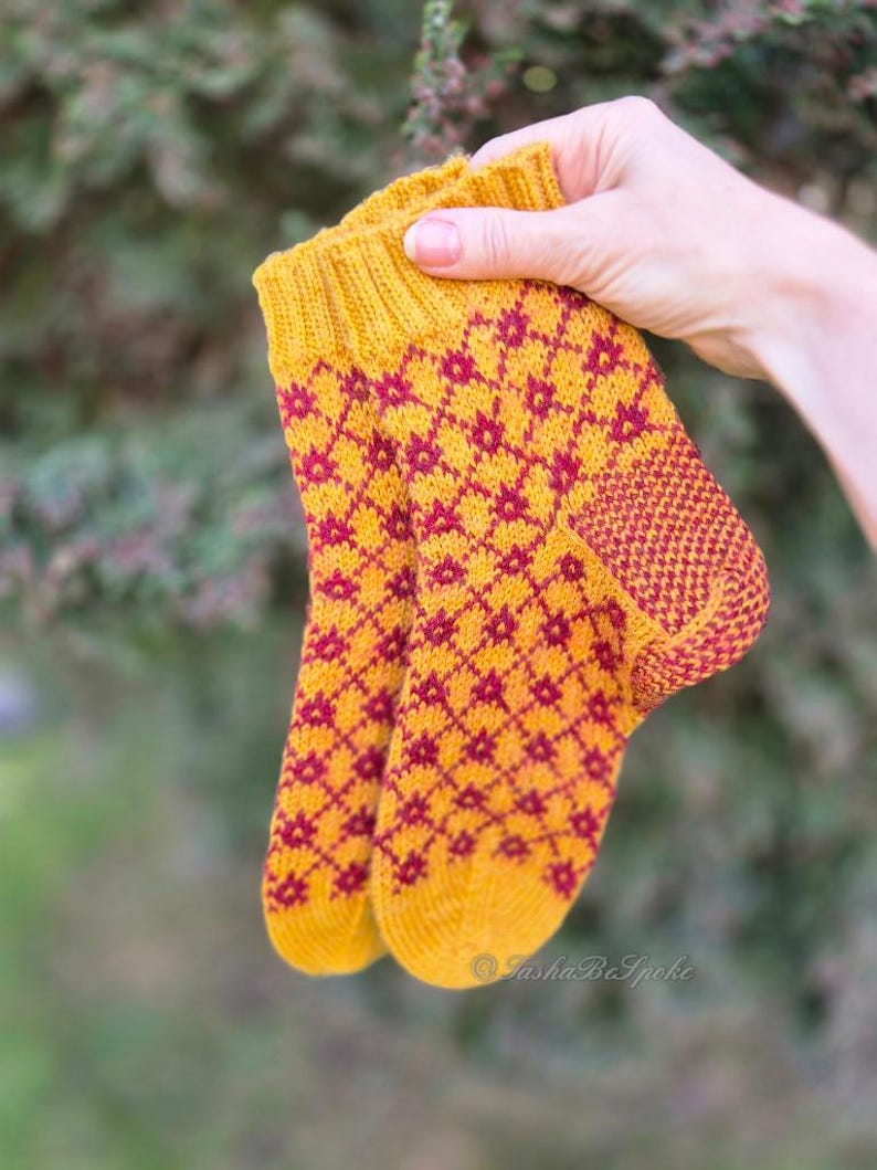 May include: A pair of knitted socks in a hand. The socks are yellow with a repeating pattern of red diamond-shaped flowers. The cuff and heel are a solid red color. The socks are held up against a blurred green background.