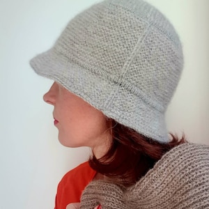 May include: A light gray knitted cloche hat with a small brim. The hat is made of a textured yarn and has a structured design. The person in the image is wearing an orange shirt and holding a brown knitted scarf.