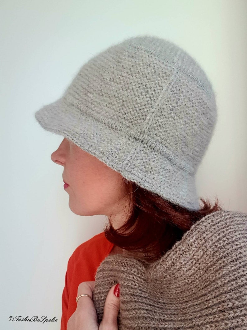 May include: A light gray knitted cloche hat with a small brim. The hat is made of a textured yarn and has a structured design. The person in the image is wearing an orange shirt and holding a brown knitted scarf.
