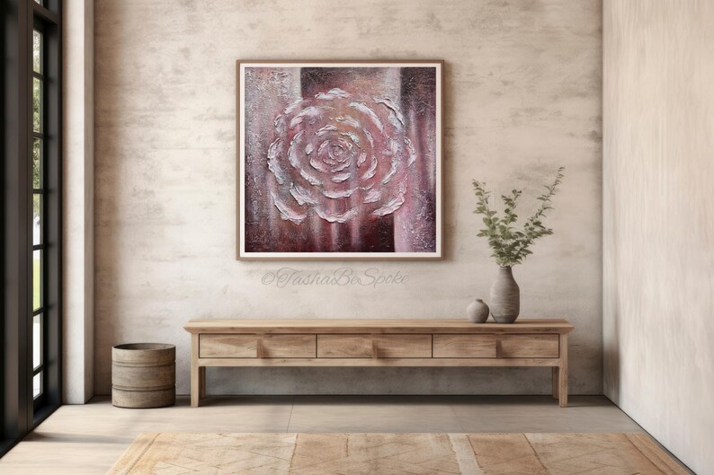May include: An abstract painting of a rose in pink and white tones is framed and displayed above a wooden console table. The artwork has textured brushstrokes, creating a 3D effect. The room has a neutral colour palette with a vase of greenery.