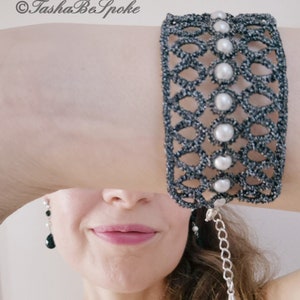 Puede incluir: Un brazalete negro y plateado con un delicado dise&ntilde;o calado y detalles de perlas blancas. El brazalete tiene una cadena con un peque&ntilde;o dije plateado colgando. &copy;TashaBeSpoke
