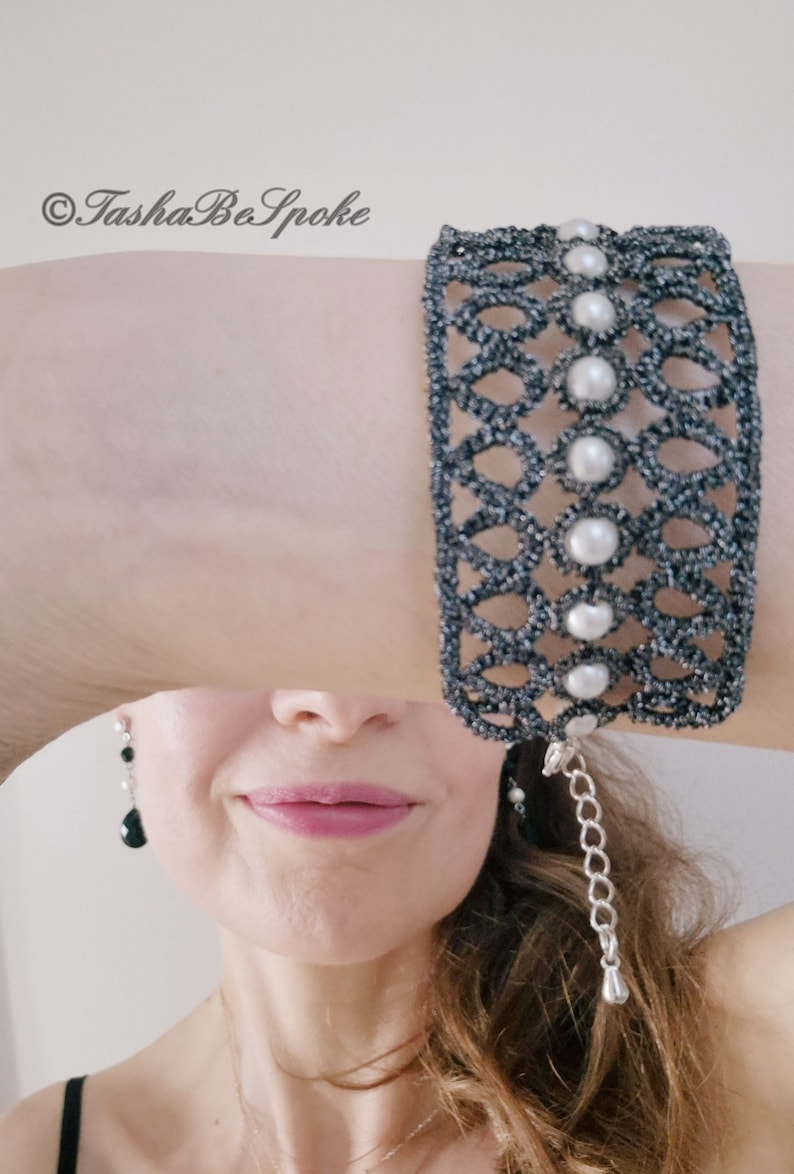 Puede incluir: Un brazalete negro y plateado con un delicado dise&ntilde;o calado y detalles de perlas blancas. El brazalete tiene una cadena con un peque&ntilde;o dije plateado colgando. &copy;TashaBeSpoke