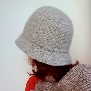 May include: A light gray knitted cloche hat with a small brim. The hat is made of a textured yarn and has a classic, rounded shape. The hat is shown from a side view.