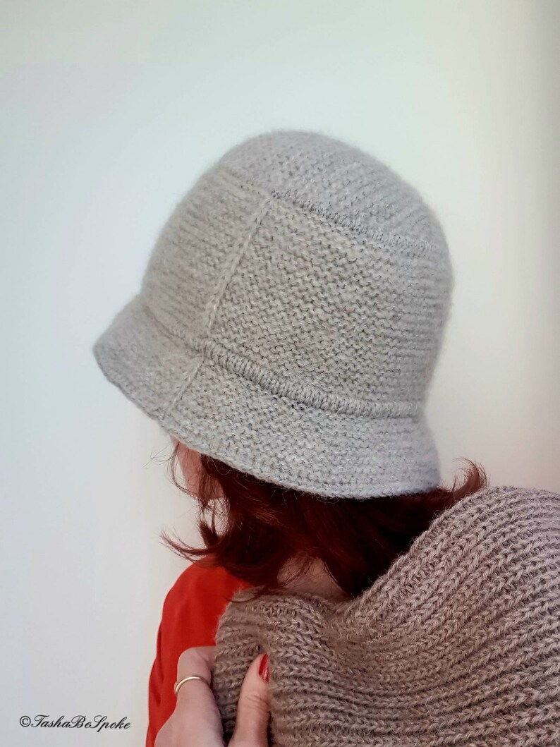 May include: A light gray knitted cloche hat with a small brim. The hat is made of a textured yarn and has a classic, rounded shape. The hat is shown from a side view.