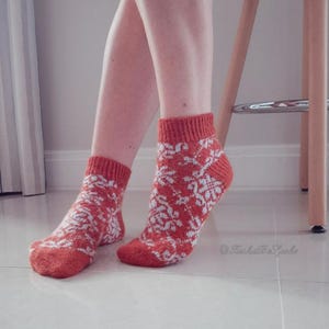 May include: A pair of ankle-length socks in a burnt orange color with a white floral pattern. The socks have a ribbed cuff and are worn on a person's feet. The background is a light-colored tiled floor and a wooden stool.