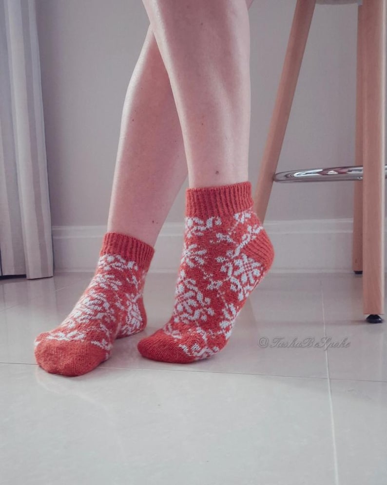 May include: A pair of ankle-length socks in a burnt orange color with a white floral pattern. The socks have a ribbed cuff and are worn on a person's feet. The background is a light-colored tiled floor and a wooden stool.