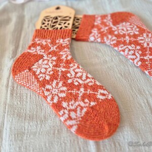 May include: A pair of orange and white knit socks with a snowflake pattern. The socks have a wooden tag attached. The socks are made of a soft, warm material, perfect for cold weather. The socks are a cozy and stylish accessory.