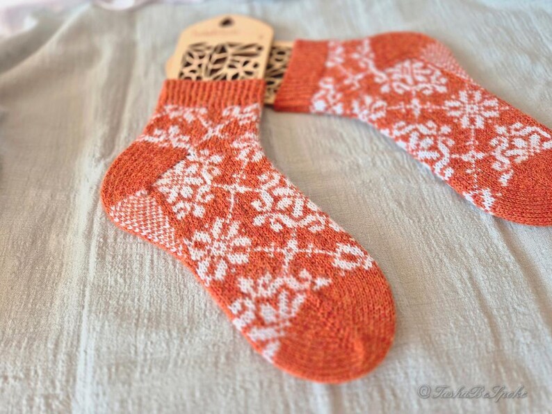 May include: A pair of orange and white knit socks with a snowflake pattern. The socks have a wooden tag attached. The socks are made of a soft, warm material, perfect for cold weather. The socks are a cozy and stylish accessory.