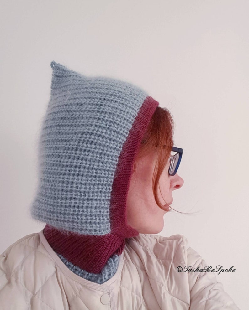 Pu&ograve; includere: Un cappuccio lavorato a maglia blu chiaro e bordeaux. Il cappuccio ha una parte superiore a punta e un bordo bordeaux intorno al viso e al collo. Il cappuccio &egrave; indossato sopra una giacca trapuntata colorata.