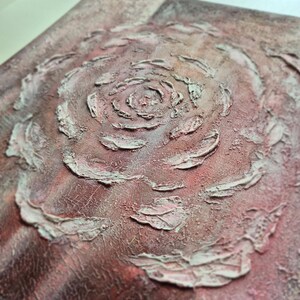 May include: Abstract artwork with a textured rose design in the centre. The piece uses shades of red, pink, and white, with a rough, tactile surface. The artwork is framed in a white border.