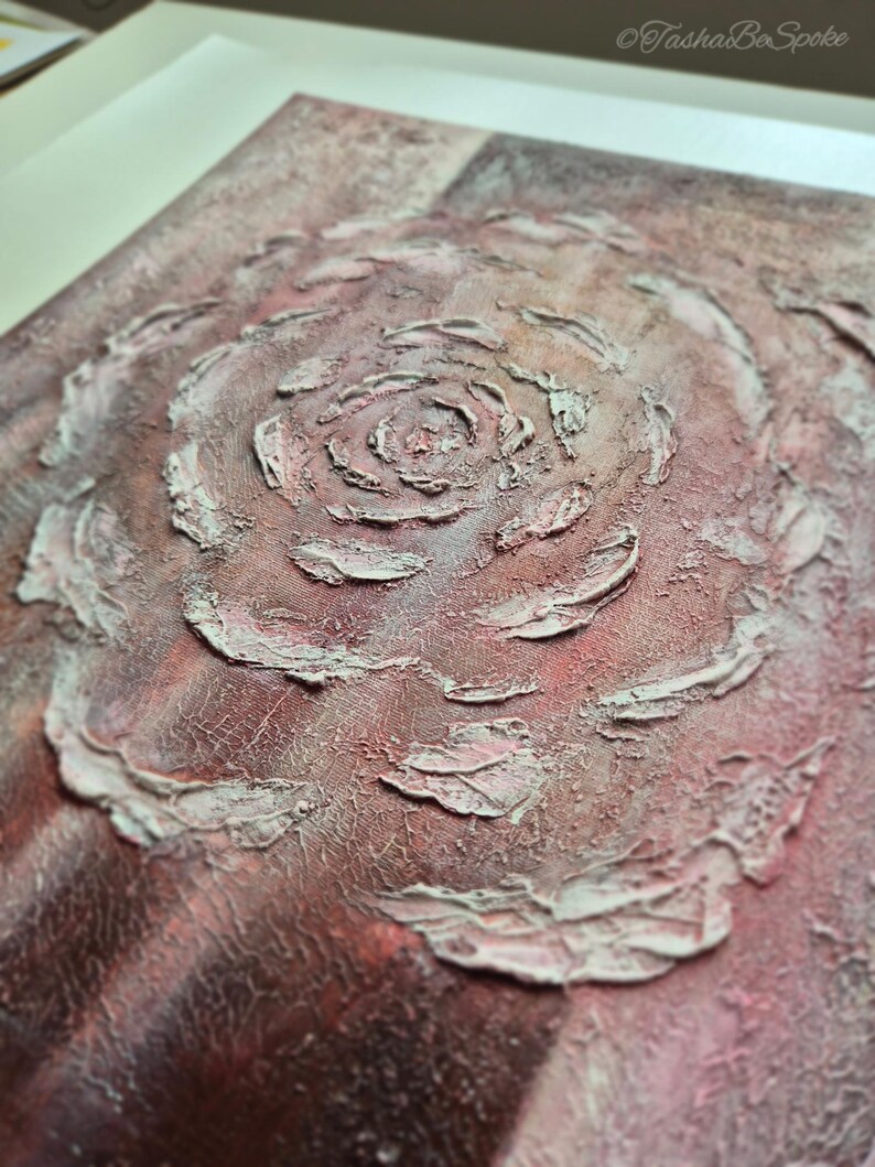 May include: Abstract artwork with a textured rose design in the centre. The piece uses shades of red, pink, and white, with a rough, tactile surface. The artwork is framed in a white border.