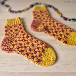 May include: A pair of knitted socks with a yellow base and a repeating maroon floral pattern. The socks have yellow cuffs, heels, and toes. A colorful beaded strand is in the background. The socks are displayed on a light wooden surface.