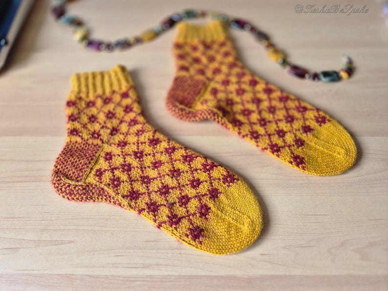 May include: A pair of knitted socks with a yellow base and a repeating maroon floral pattern. The socks have yellow cuffs, heels, and toes. A colorful beaded strand is in the background. The socks are displayed on a light wooden surface.
