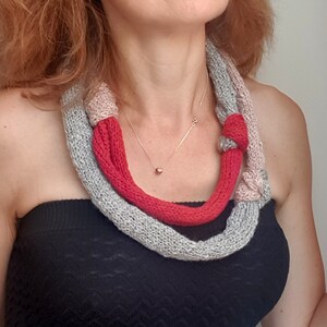 May include: A knitted necklace in shades of gray, pink, and red. The necklace is wrapped around the neck and features a knotted design. A gold necklace is also visible.