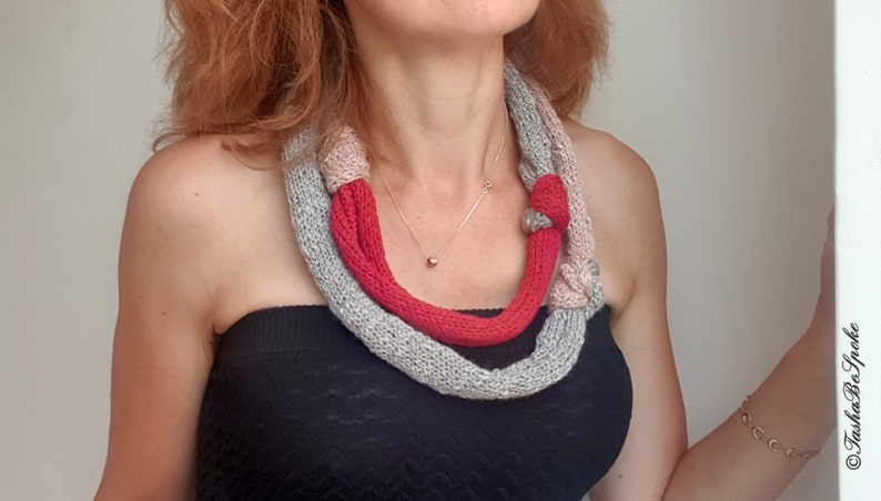 May include: A knitted necklace in shades of gray, pink, and red. The necklace is wrapped around the neck and features a knotted design. A gold necklace is also visible.