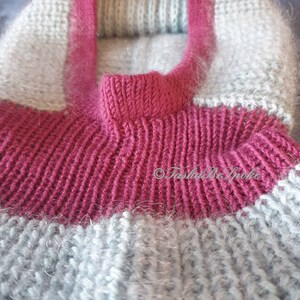 Pu&ograve; includere: Primo piano di un maglione lavorato a mano a maglia nei toni del grigio e magenta. Il maglione presenta un colletto magenta a coste e una fascia abbinata sul petto. Il corpo del maglione &egrave; di colore grigio chiaro.