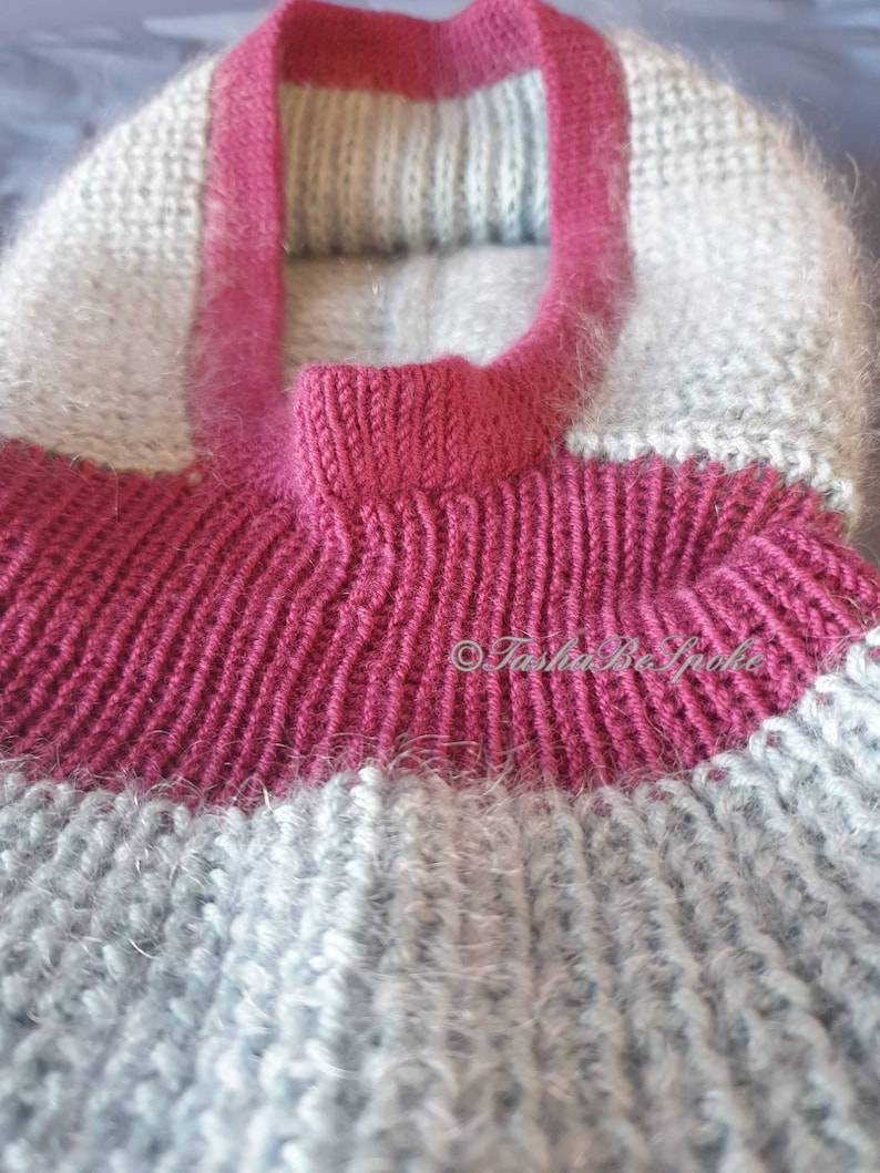 Pu&ograve; includere: Primo piano di un maglione lavorato a mano a maglia nei toni del grigio e magenta. Il maglione presenta un colletto magenta a coste e una fascia abbinata sul petto. Il corpo del maglione &egrave; di colore grigio chiaro.