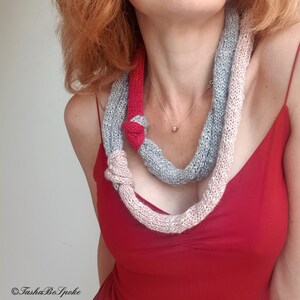 May include: A knitted scarf in shades of gray, pink, and red, knotted in two places. The scarf is worn over a red dress. The scarf is a unique accessory.