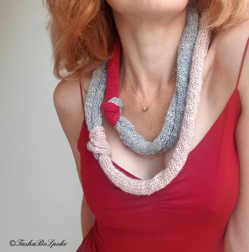 May include: A knitted scarf in shades of gray, pink, and red, knotted in two places. The scarf is worn over a red dress. The scarf is a unique accessory.