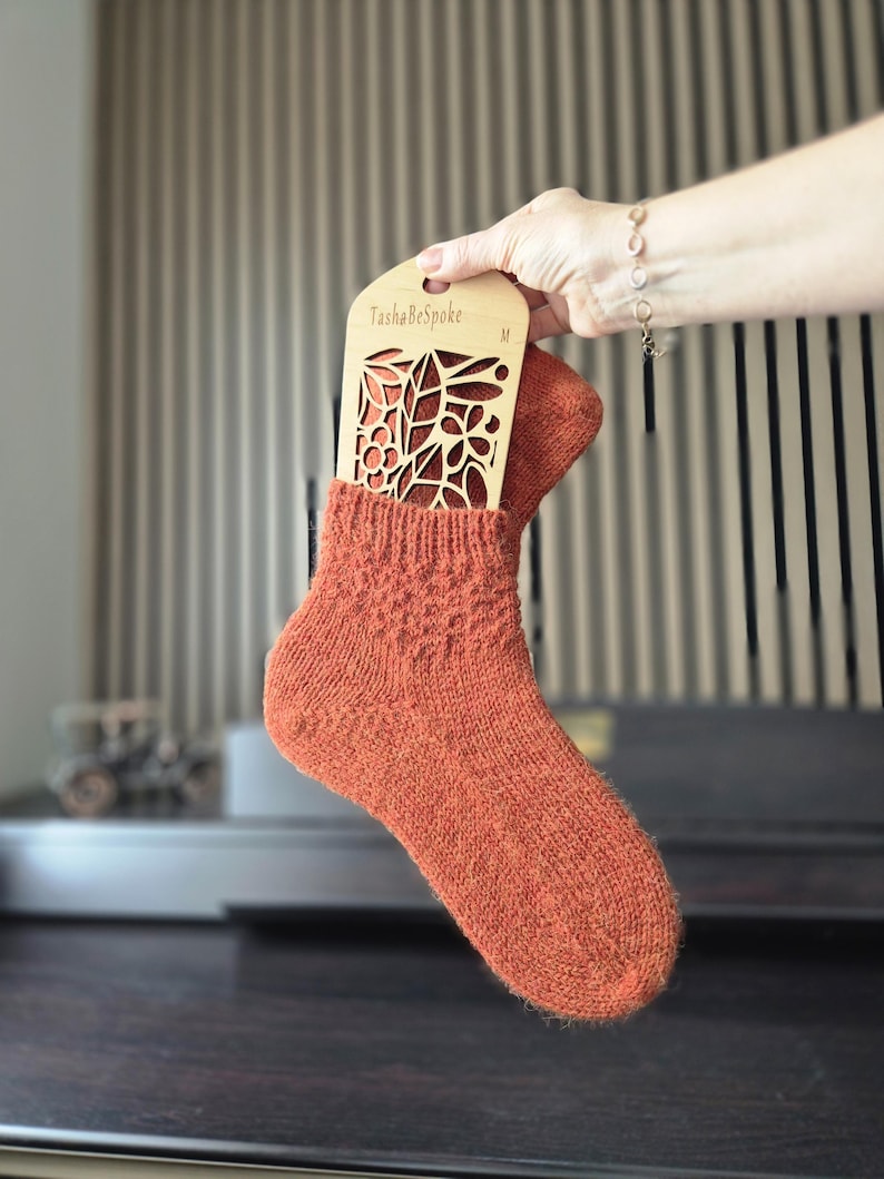 May include: An orange knitted sock is held with a wooden sock blocker. The sock blocker has a floral design cut out and the text "TashaBeSpoke" is visible. The sock is a medium size.
