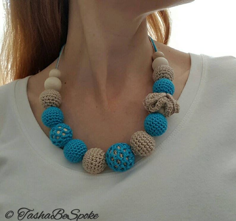Op de afbeelding: Een handgemaakte ketting met een verscheidenheid aan kralen. De ketting bevat lichtblauwe gehaakte kralen, beige gehaakte kralen en natuurlijke houten kralen. De ketting is geregen op een lichtblauw koord.