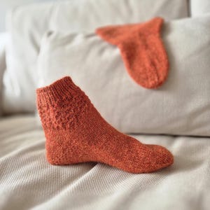 May include: An orange knitted sock with a textured pattern on the cuff, resting on a neutral-toned surface. A matching sock is in the background. The socks appear to be handmade, showcasing a cozy, warm aesthetic.