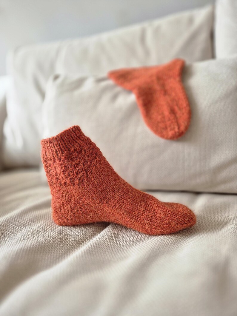 May include: An orange knitted sock with a textured pattern on the cuff, resting on a neutral-toned surface. A matching sock is in the background. The socks appear to be handmade, showcasing a cozy, warm aesthetic.
