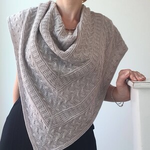May include: A light brown knitted shawl with a cable knit pattern. The shawl is draped around a person's neck and shoulders.