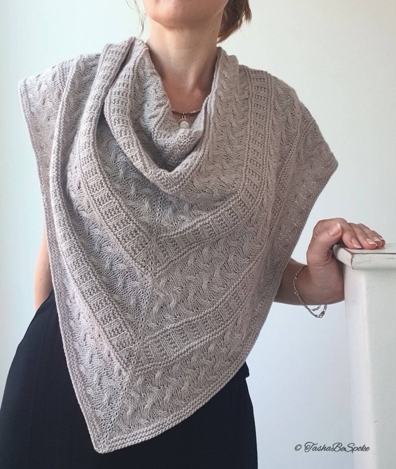May include: A light brown knitted shawl with a cable knit pattern. The shawl is draped around a person's neck and shoulders.