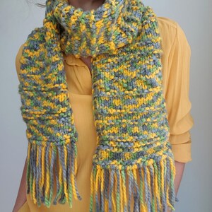 May include: A knitted scarf with yellow, green, and gray stripes and fringe. The scarf is worn by a person wearing a yellow shirt.