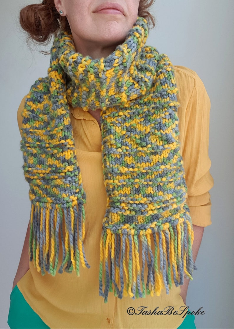 May include: A knitted scarf with yellow, green, and gray stripes and fringe. The scarf is worn by a person wearing a yellow shirt.