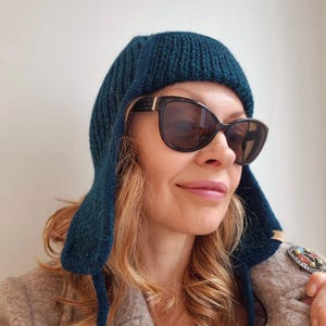 K&ouml;nnte beinhalten: Eine t&uuml;rkisblaue Strickm&uuml;tze mit Ohrenklappen. Die M&uuml;tze ist &uuml;ber die Stirn gefaltet und hat eine gerippte Struktur. Die Person, die die M&uuml;tze tr&auml;gt, tr&auml;gt auch eine Sonnenbrille.