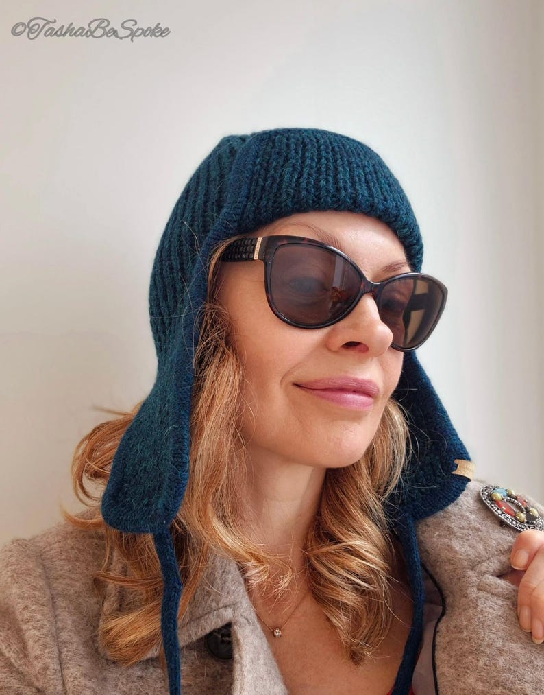 K&ouml;nnte beinhalten: Eine t&uuml;rkisblaue Strickm&uuml;tze mit Ohrenklappen. Die M&uuml;tze ist &uuml;ber die Stirn gefaltet und hat eine gerippte Struktur. Die Person, die die M&uuml;tze tr&auml;gt, tr&auml;gt auch eine Sonnenbrille.