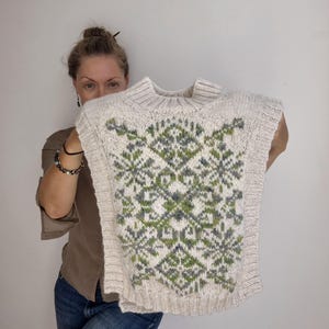 May include: A sleeveless knit sweater vest in an off-white color featuring a prominent geometric and floral pattern in shades of sage green and dark gray. The garment has a ribbed mock neck collar, ribbed armholes, and a ribbed hemline.