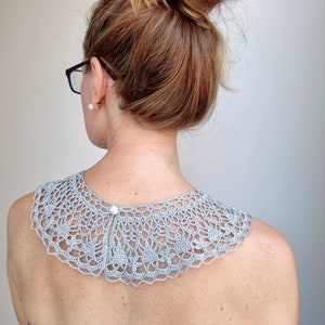 May include: A light gray crocheted collar with a pearl button closure. The collar is worn by a person with their hair pulled back in a bun.