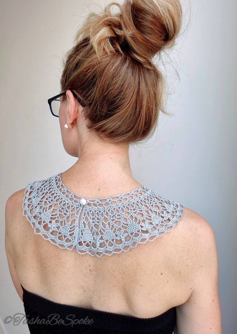 May include: A light gray crocheted collar with a pearl button closure. The collar is worn by a person with their hair pulled back in a bun.
