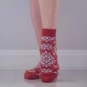 May include: Red and white patterned socks. The socks feature a repeating snowflake design in white against a red background. The socks are knit and have a ribbed cuff. The socks are worn on a person's feet.