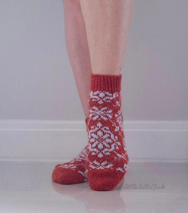 May include: Red and white patterned socks. The socks feature a repeating snowflake design in white against a red background. The socks are knit and have a ribbed cuff. The socks are worn on a person's feet.
