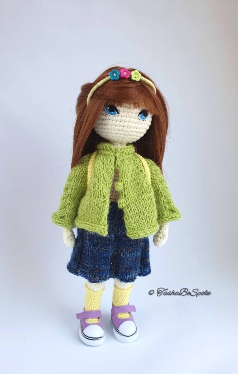 Pu&ograve; includere: Una bambola all'uncinetto con lunghi capelli castani, che indossa un cardigan verde, una gonna blu, calzini gialli e scarpe viola. La bambola ha gli occhi azzurri e indossa una fascia per capelli con fiori colorati.
