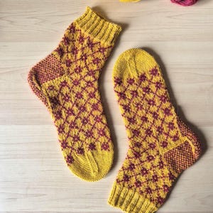 May include: A pair of hand-knitted socks in a diamond pattern. The socks are primarily yellow with a burgundy diamond pattern. The heels and toes are burgundy. Two balls of yarn, one yellow and one burgundy, are in the background.