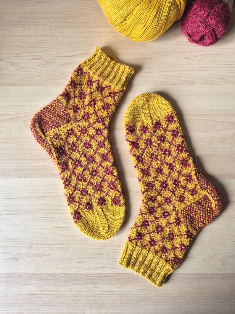 May include: A pair of hand-knitted socks in a diamond pattern. The socks are primarily yellow with a burgundy diamond pattern. The heels and toes are burgundy. Two balls of yarn, one yellow and one burgundy, are in the background.