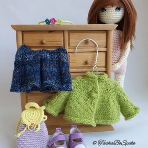 Pu&ograve; includere: Una bambola all'uncinetto con capelli castani e occhi azzurri che indossa un vestito rosa. La bambola &egrave; in piedi accanto a una cassettiera di legno con cassetti. Ci sono diversi oggetti all'uncinetto sulla cassettiera, tra cui un cardigan verde, una gonna blu, una borsa viola e un paio di scarpe viola. Il testo "Tasha Be Spoke" &egrave; visibile nell'angolo in basso a destra dell'immagine.