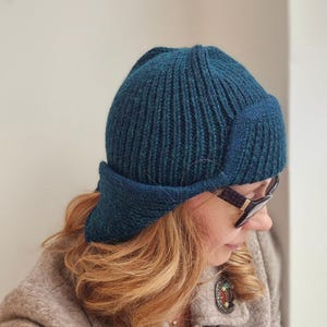K&ouml;nnte beinhalten: Eine tealblaue Strickm&uuml;tze mit breitem Rand und Ohrenklappen. Die M&uuml;tze ist aus weichem, strukturiertem Garn gefertigt und hat ein geripptes Design.