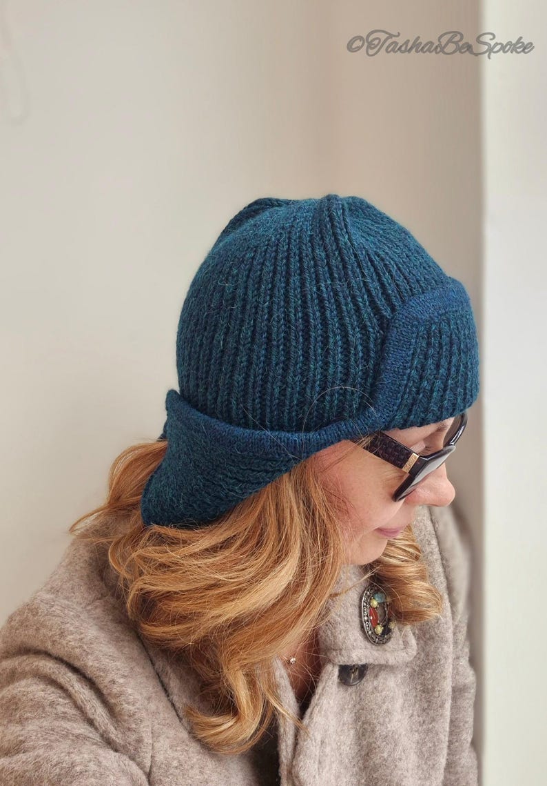 K&ouml;nnte beinhalten: Eine tealblaue Strickm&uuml;tze mit breitem Rand und Ohrenklappen. Die M&uuml;tze ist aus weichem, strukturiertem Garn gefertigt und hat ein geripptes Design.