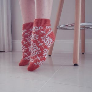 May include: A pair of orange and white patterned socks. The socks have a snowflake design and are worn on the feet. The socks are made of a knitted material. The image is taken indoors.
